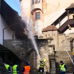 Fotografia zachytávajúca príslušníka hasičského zboru v procese ochladzovania strešnej drevenej krytiny prúdnou hadicou.